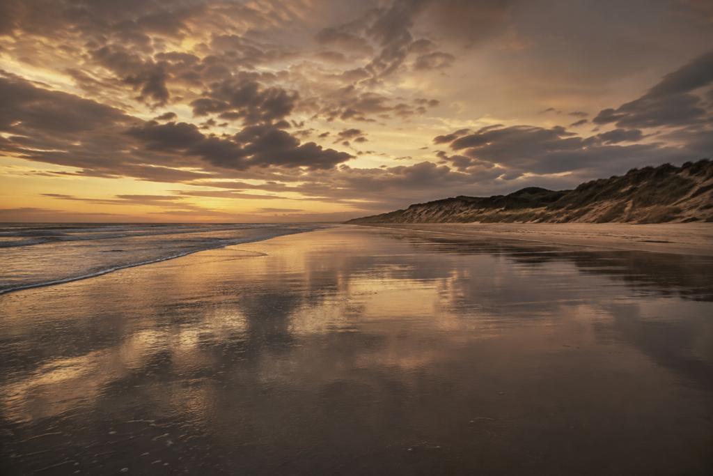 Wide view of a quiet Normandy D-Day landing beach at golden hour with wet sand and dunes.